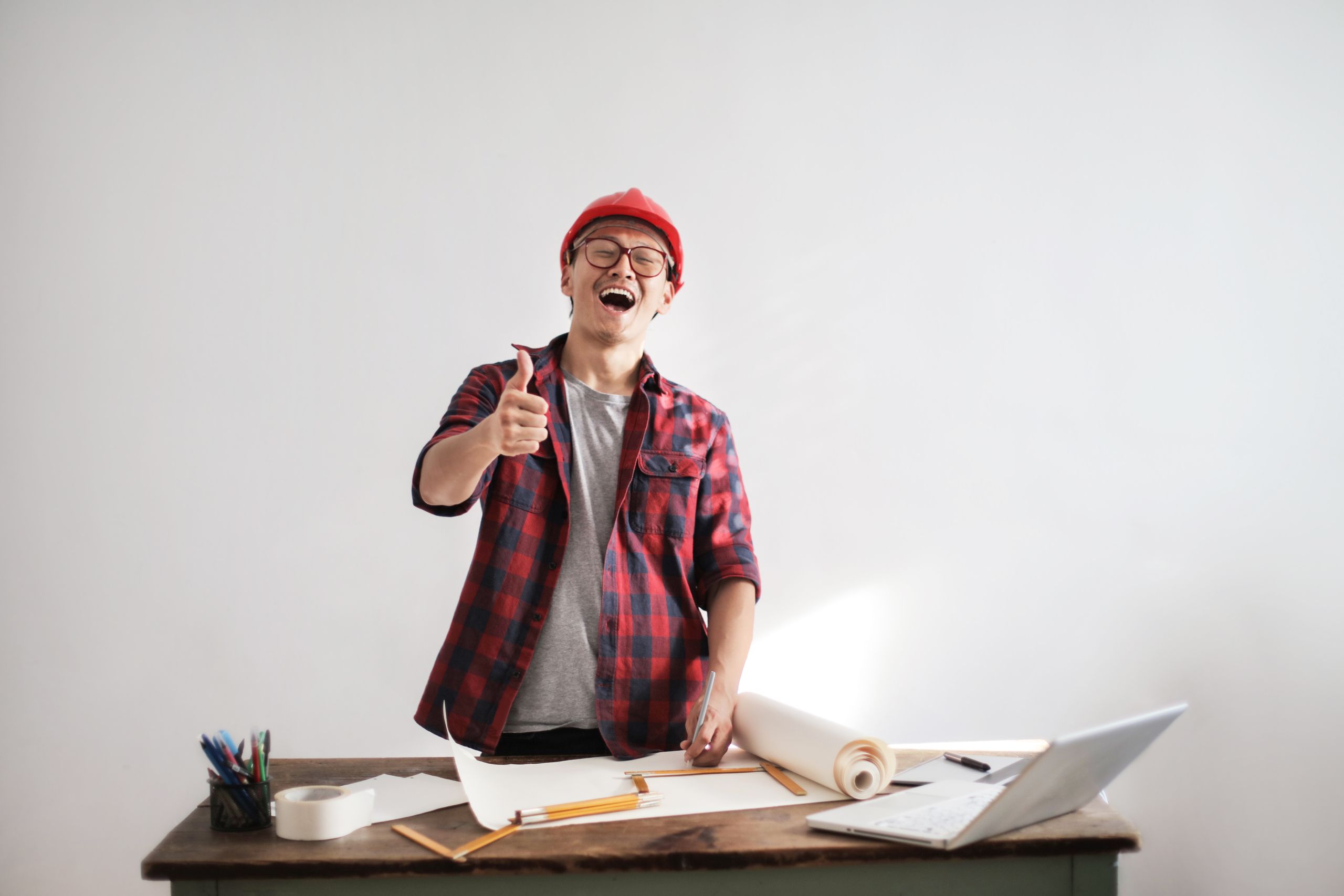 Happy young man in plaid shirt and hardhat showing thumb up standing at table with draw and laptop on white background