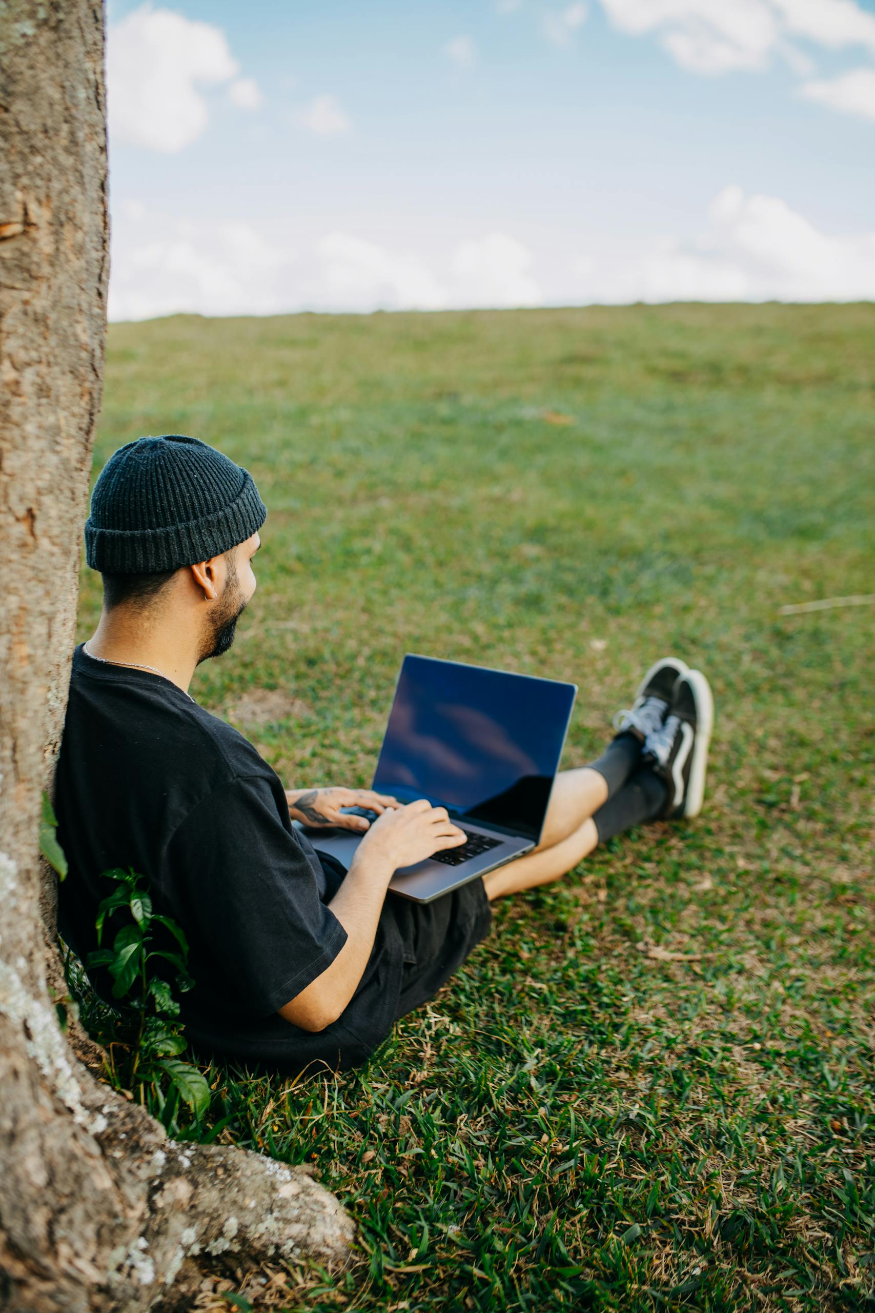 Man sitting against a tree, working on a laptop in a grassy field. Perfect for outdoor work themes.