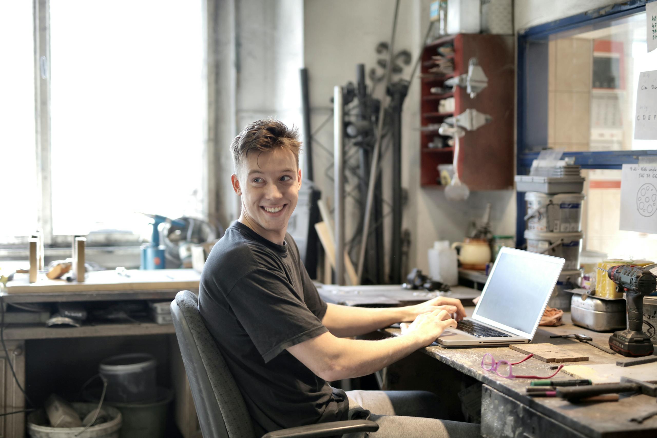 Young man smiling while using laptop in a creative workspace with tools around.