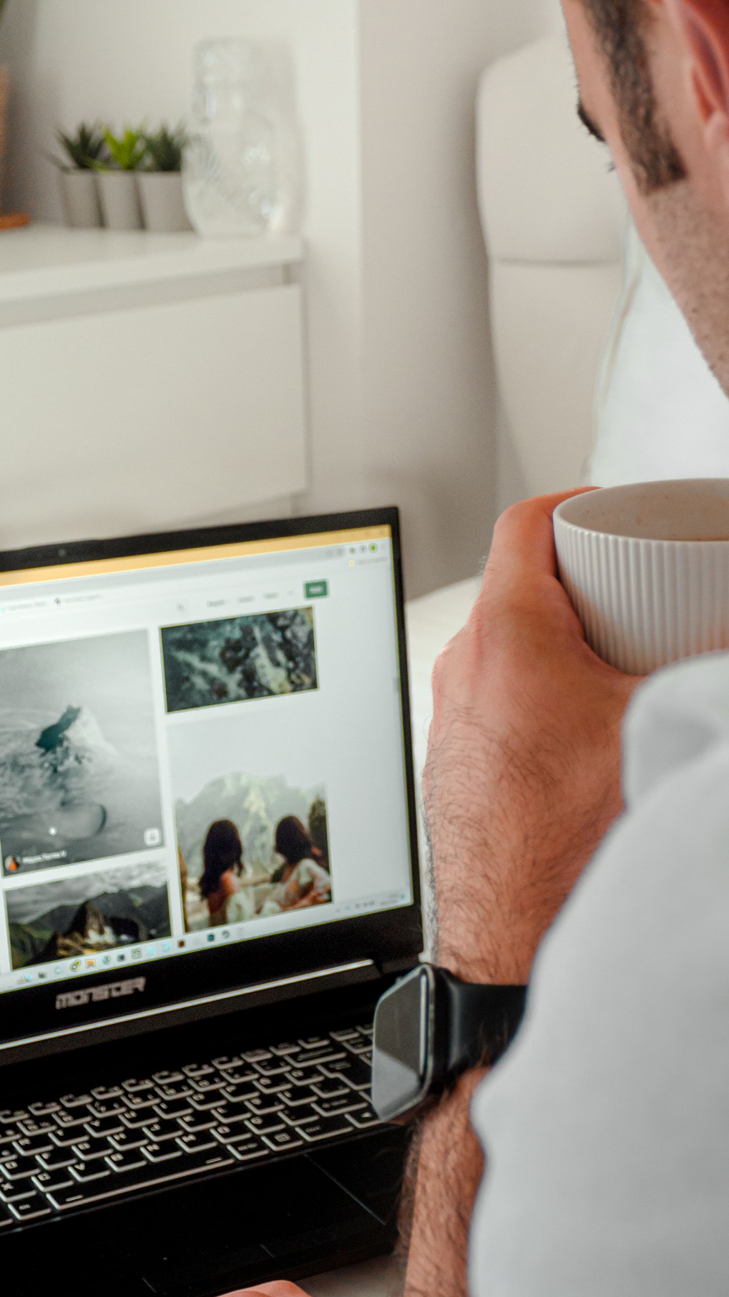 A man in casual attire holds a coffee cup while browsing images on a laptop in a cozy indoor setting.