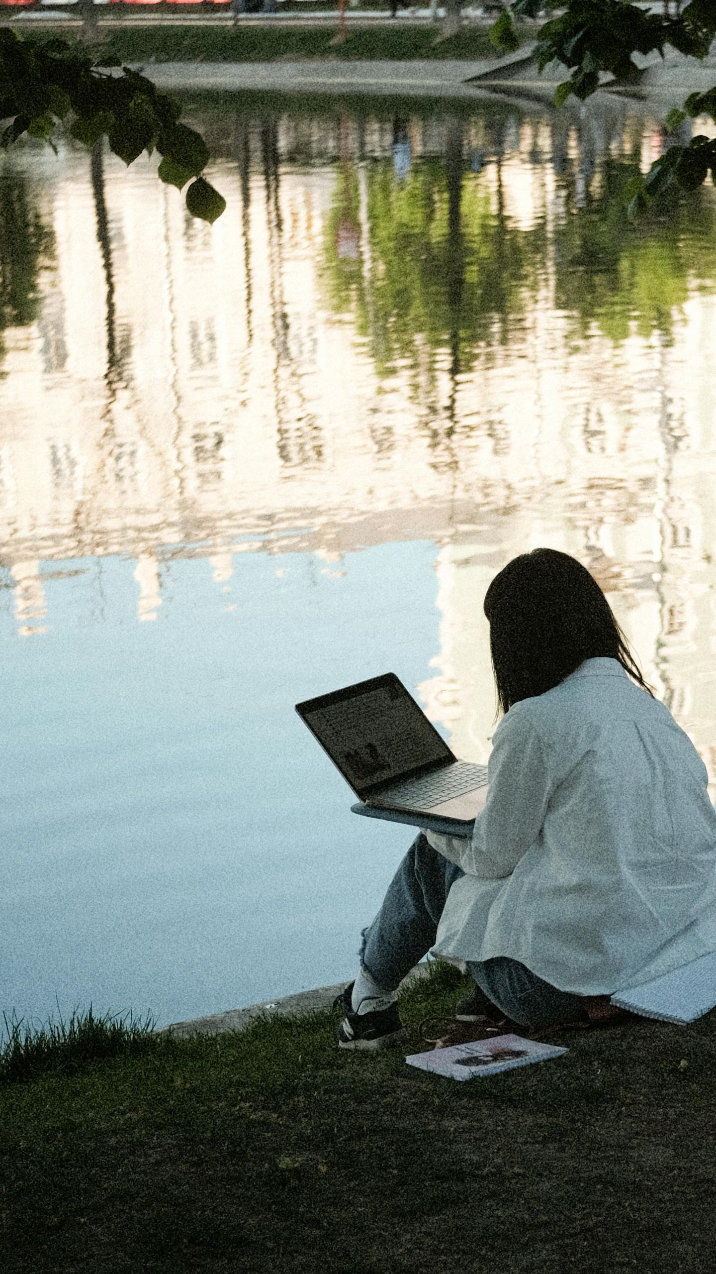 A woman working remotely on a laptop by a pond in a peaceful park setting, reflecting calm urban architecture.