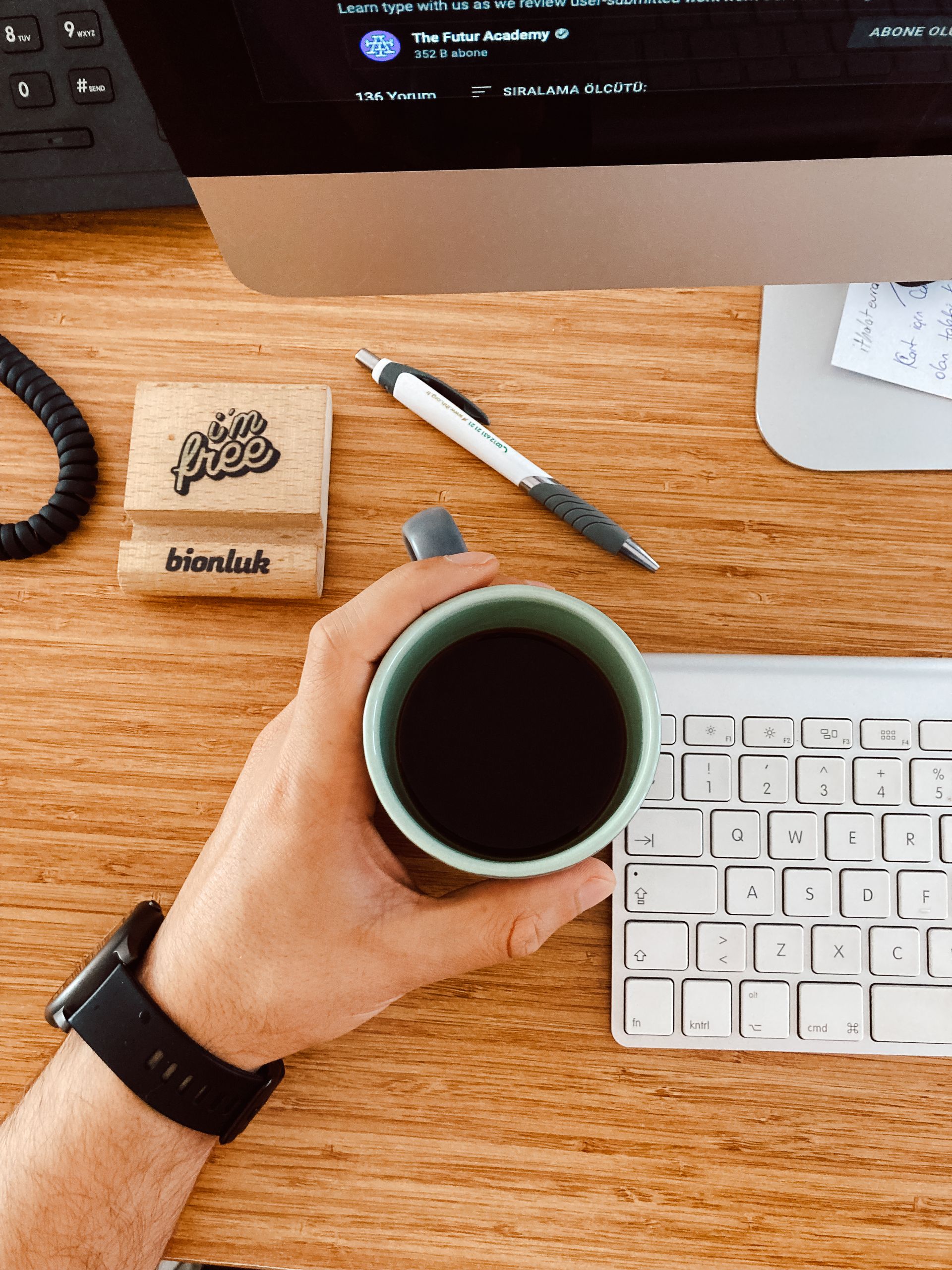 Close-up of a hand holding a coffee cup on a wooden desk with computer, keyboard, and accessories.